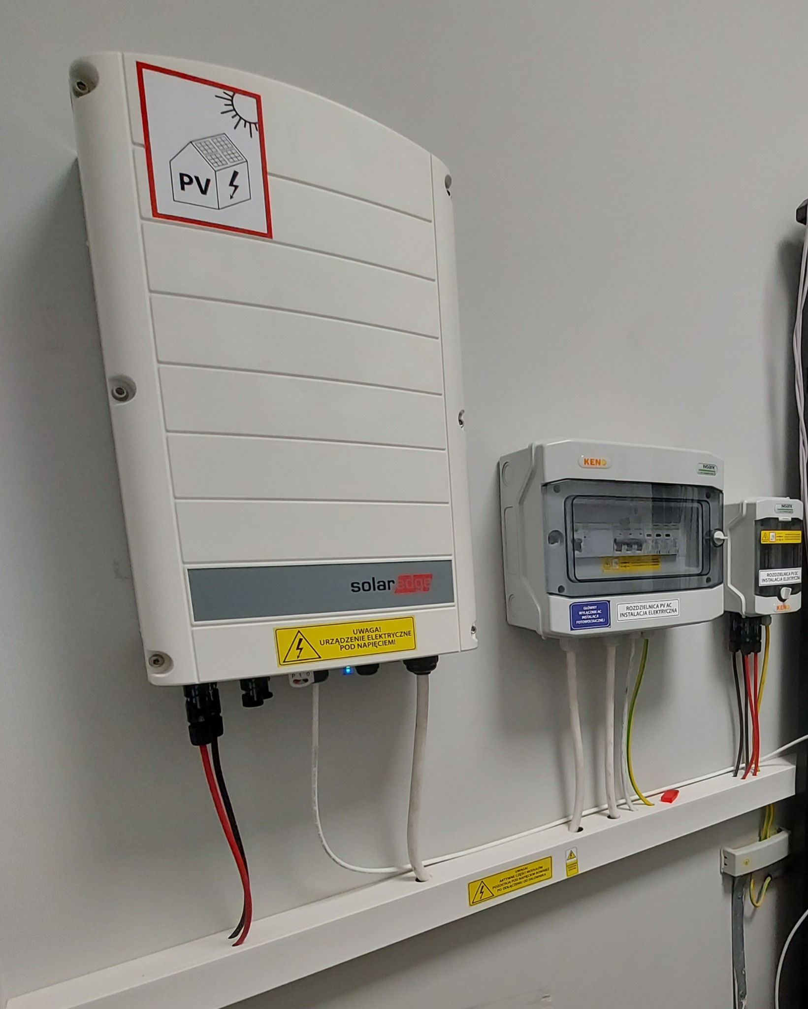 Biały inwerter SolarEdge z naklejką PV i żółtą etykietą ostrzegawczą pod napięciem, obok rozdzielnica PV AC w białej obudowie KENO z widocznymi bezpiecznikami, zamontowane na białej ścianie...