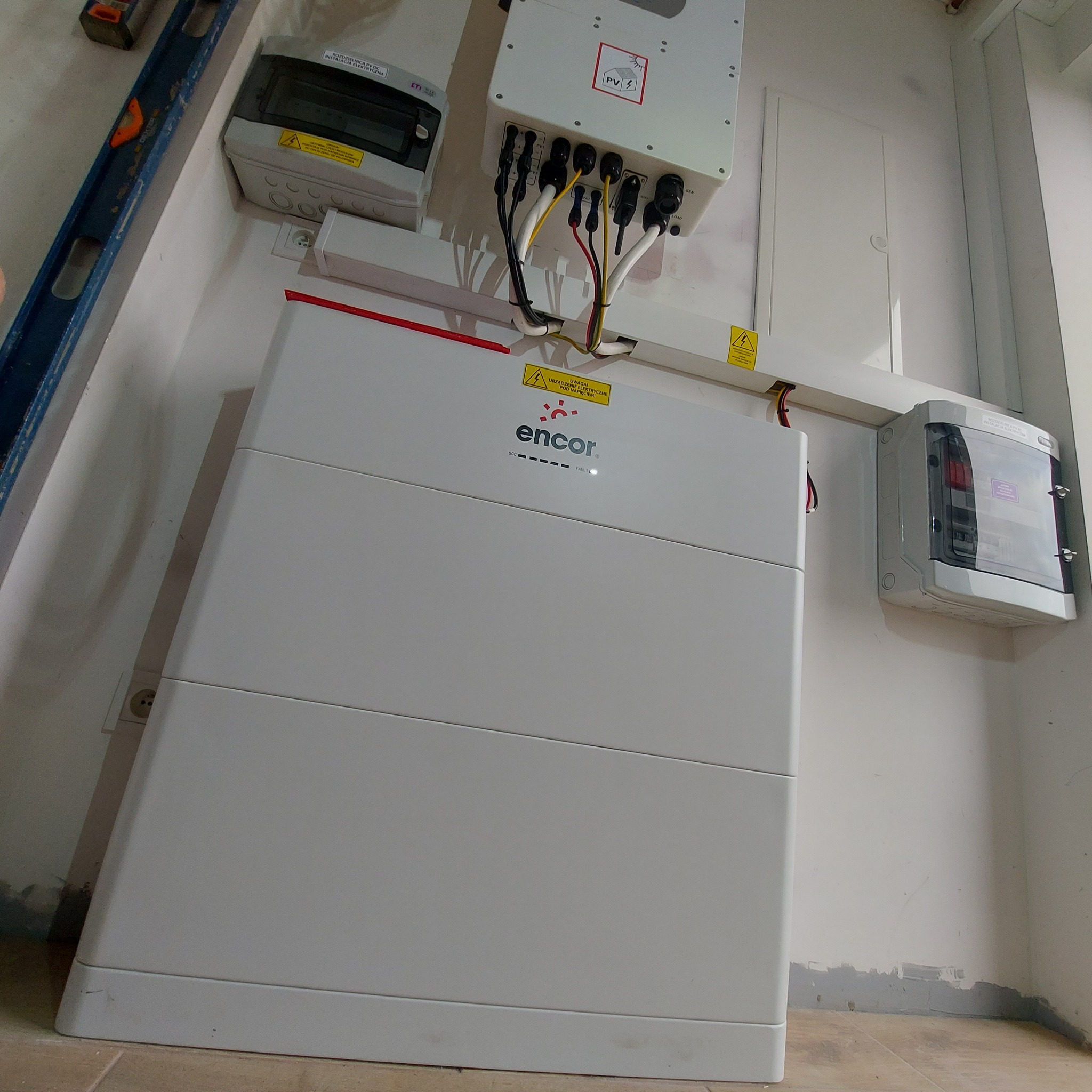 Biały akumulator energii Encor na ścianie z widocznymi przewodami, skrzynkami elektrycznymi i poziomicą, ukazujący instalację w pomieszczeniu.