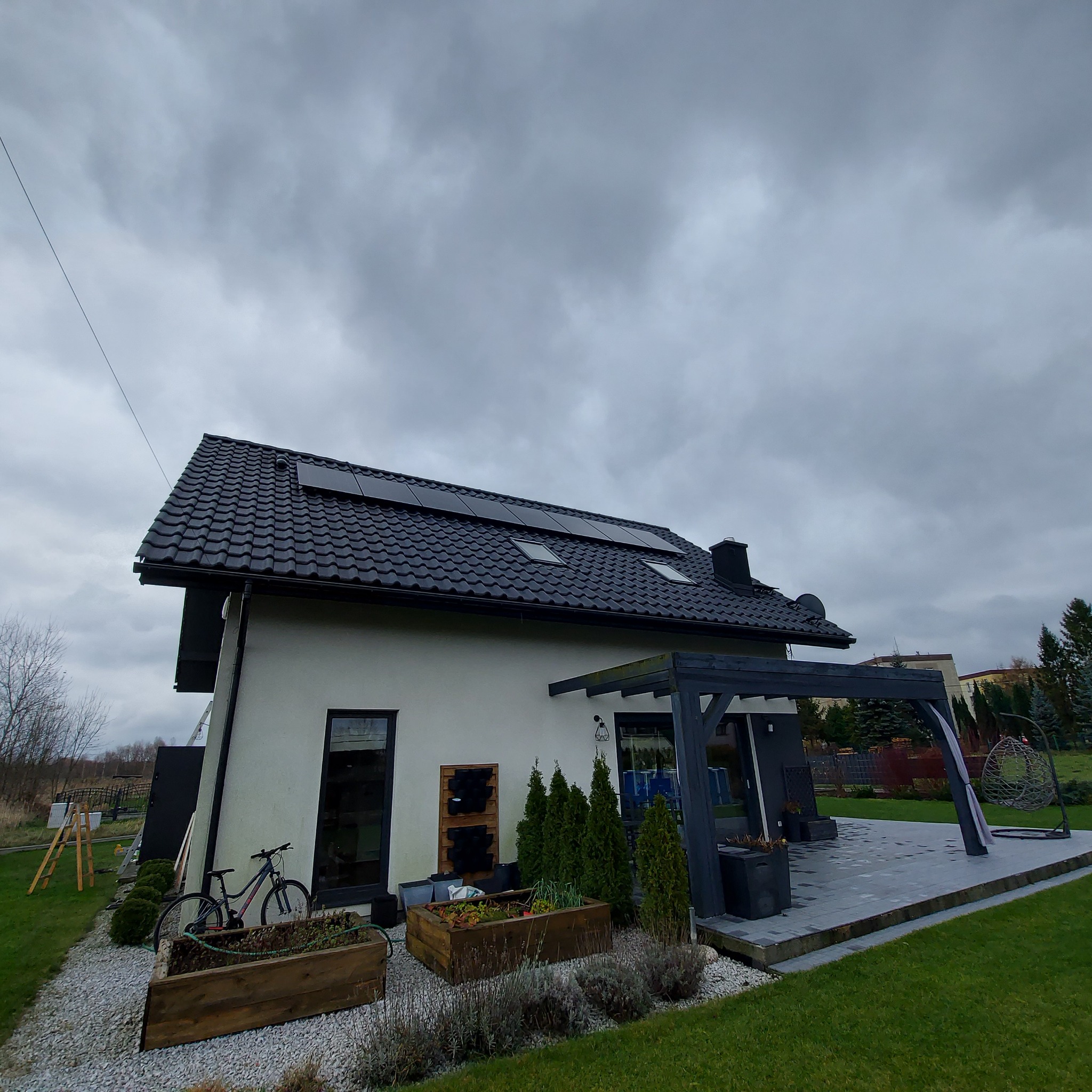 Nowoczesny dom jednorodzinny z ciemną dachówką i panelami na dachu, widok z ogrodu w pochmurny dzień, z rowerem opartym o ścianę i drewnianymi skrzyniami na rośliny.