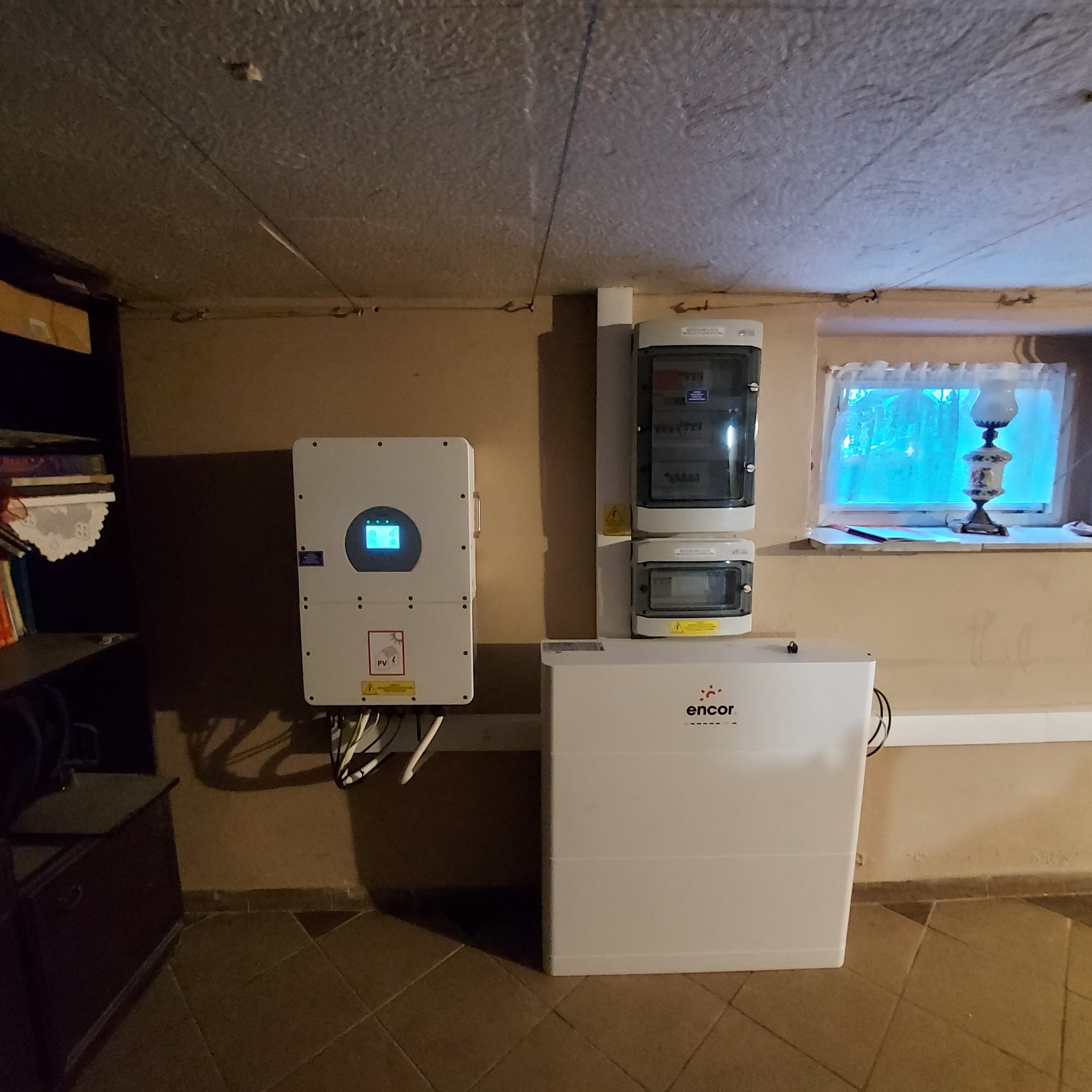Biały inwerter z wyświetlaczem, skrzynka bezpiecznikowa i magazyn energii Encor w pomieszczeniu z oknem i lampą na parapecie.