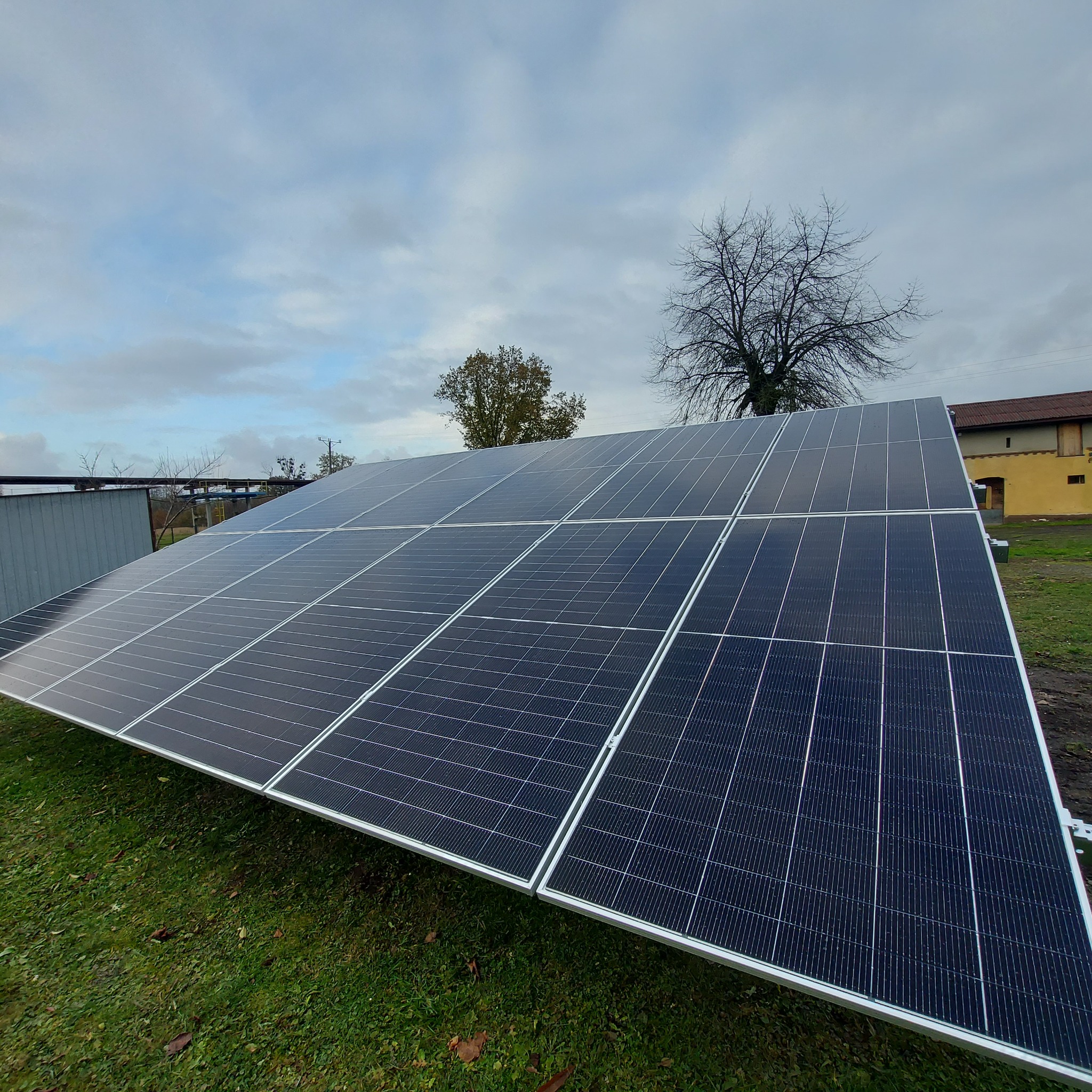 Panel słoneczny ustawiony na trawie w pochmurny dzień, widoczne drzewa w tle i fragment budynku gospodarczego.