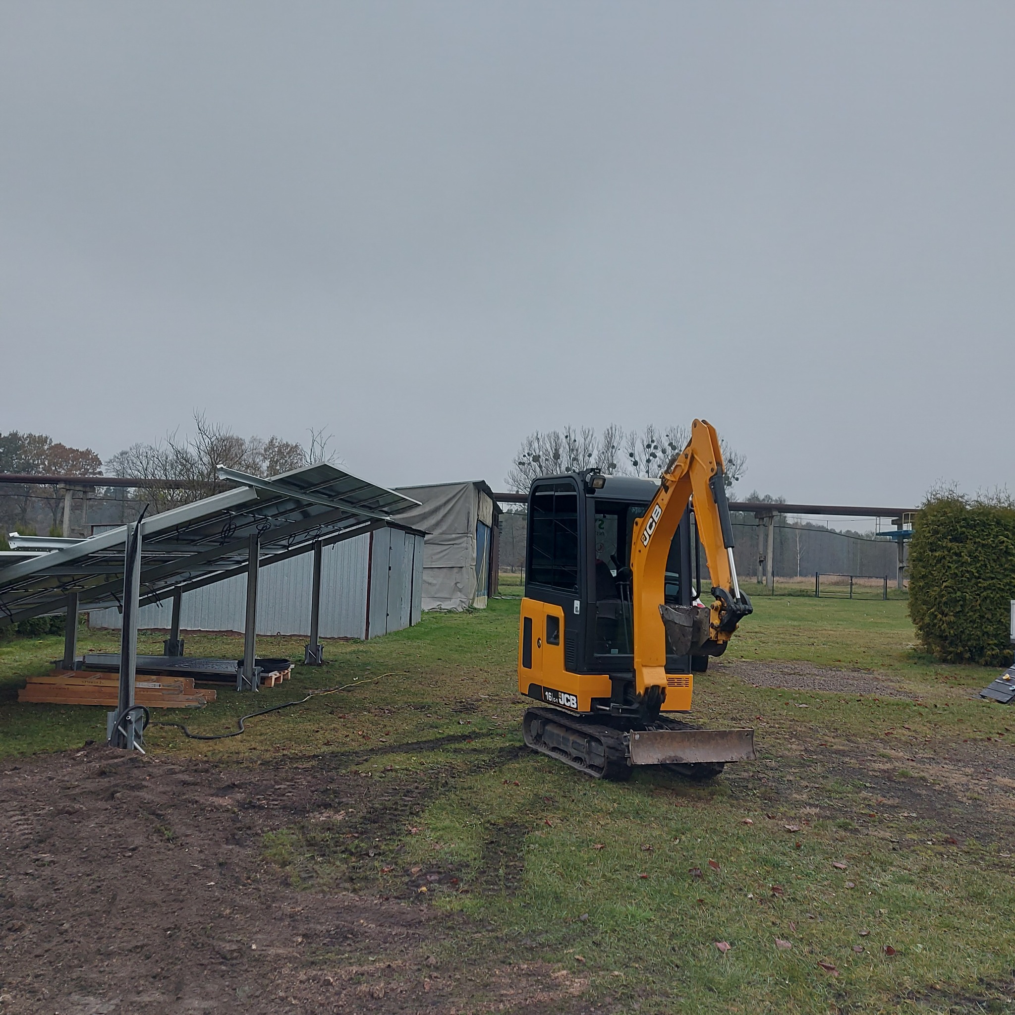 Żółta minikoparka JCB na trawiastym terenie, w tle częściowo zamontowane panele słoneczne i budynki gospodarcze w pochmurny dzień.