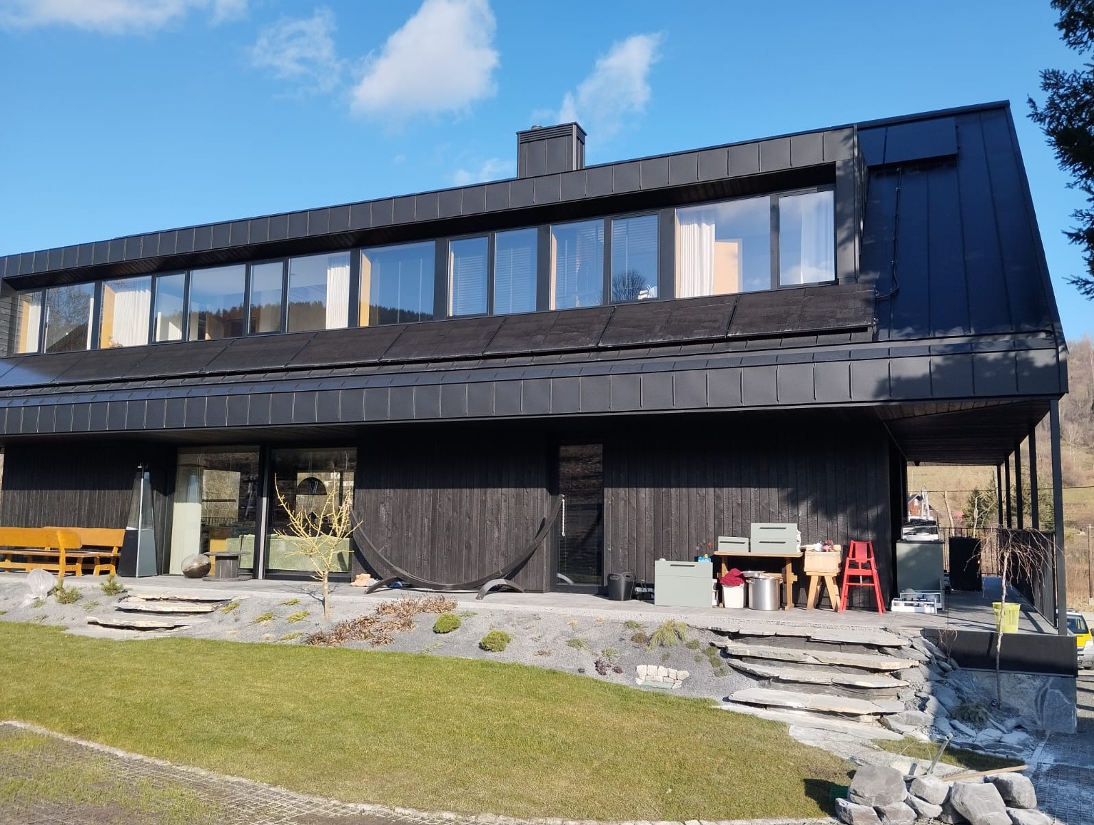 Nowoczesny dom z ciemnego drewna z panelami słonecznymi na dachu, widoczne z zewnątrz, z elementami wykończeniowymi i narzędziami na tarasie.