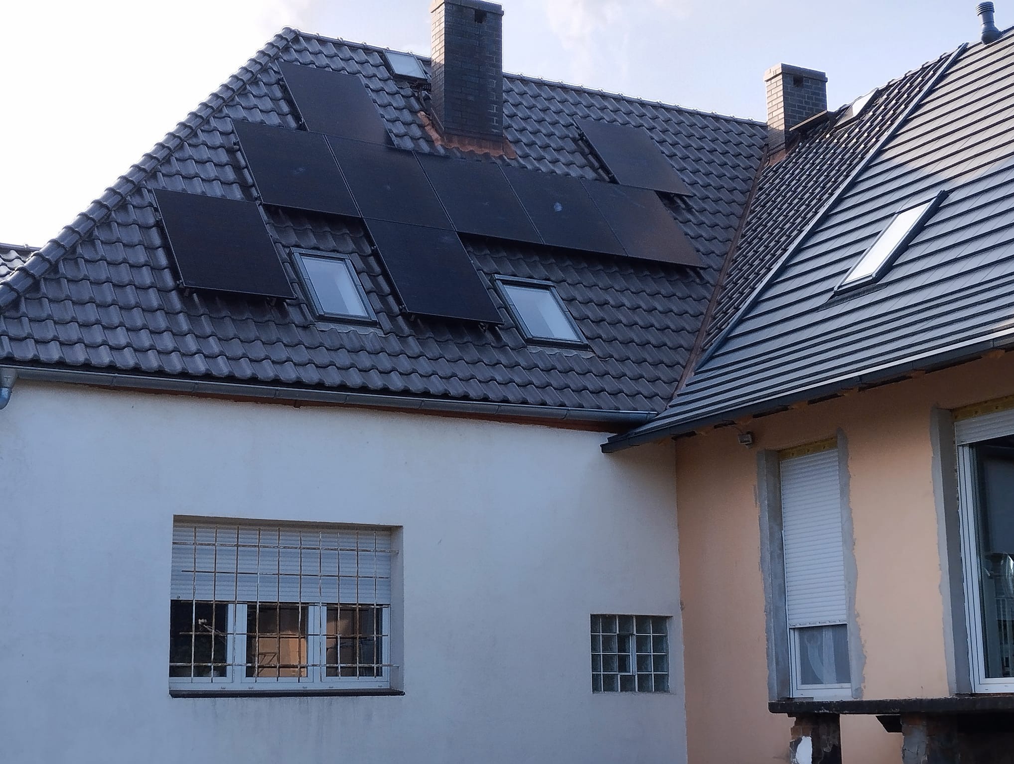 Ciemne panele słoneczne na szarym dachu dwuspadowym z oknami dachowymi i kominem, widok z dołu na tle jasnego nieba.