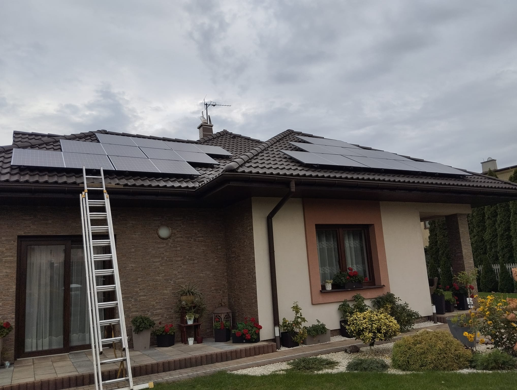 Dom jednorodzinny z brązowym dachem, na którym zamontowano panele słoneczne; aluminiowa drabina oparta o ścianę budynku.