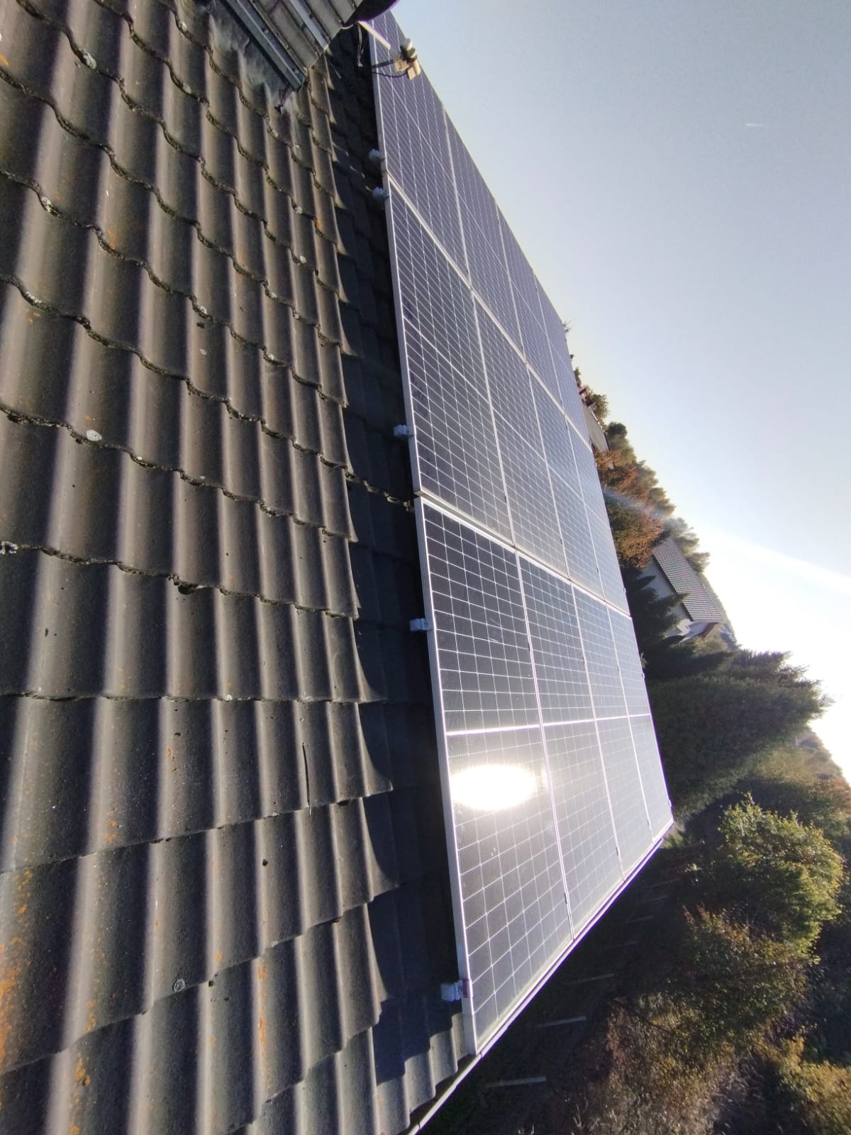 Panel słoneczny zamontowany na dachu pokrytym ciemną dachówką, widoczne mocowania i refleksy słoneczne.