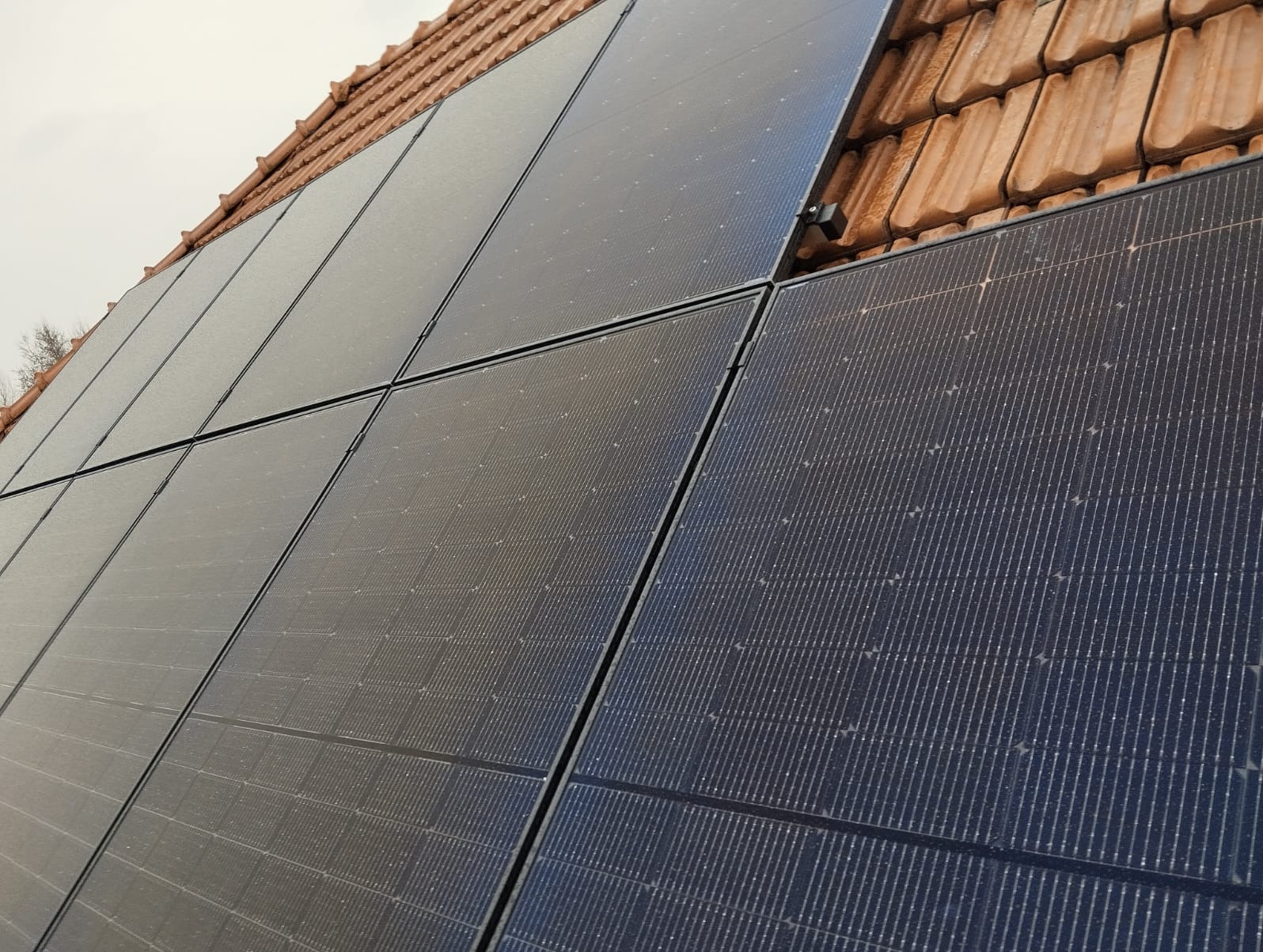 Panele słoneczne na dachu z pomarańczowej dachówki, widoczne łączenia paneli i drobne zabrudzenia na ich powierzchni.