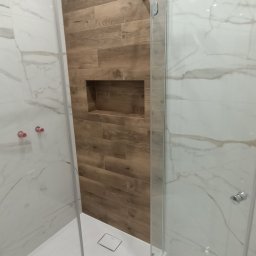 HEDAN - Nowoczesna kabina prysznicowa typu walk-in z drewnopodobną ścianą z wnęką, białe marmurowe płytki na pozostałych ścianach, odpływ liniowy w posadzce.