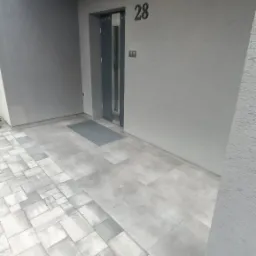 Nowoczesne wejście do domu z szarymi płytami chodnikowymi i elewacją w odcieniach szarości. Widoczne drzwi wejściowe, numer domu 28 i kamera monitoringu.