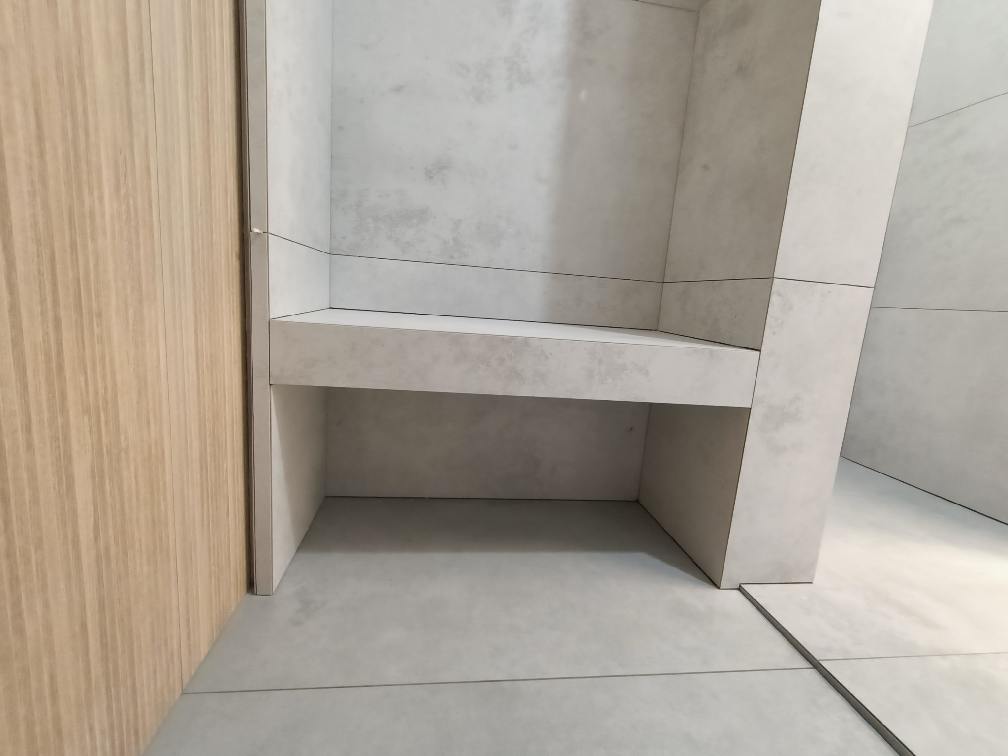 Minimalistyczna łazienka z zabudową półki i ścian wykończonych jasnoszarą, imitującą beton, płytką wielkoformatową, widoczna drewniana okładzina ściany.