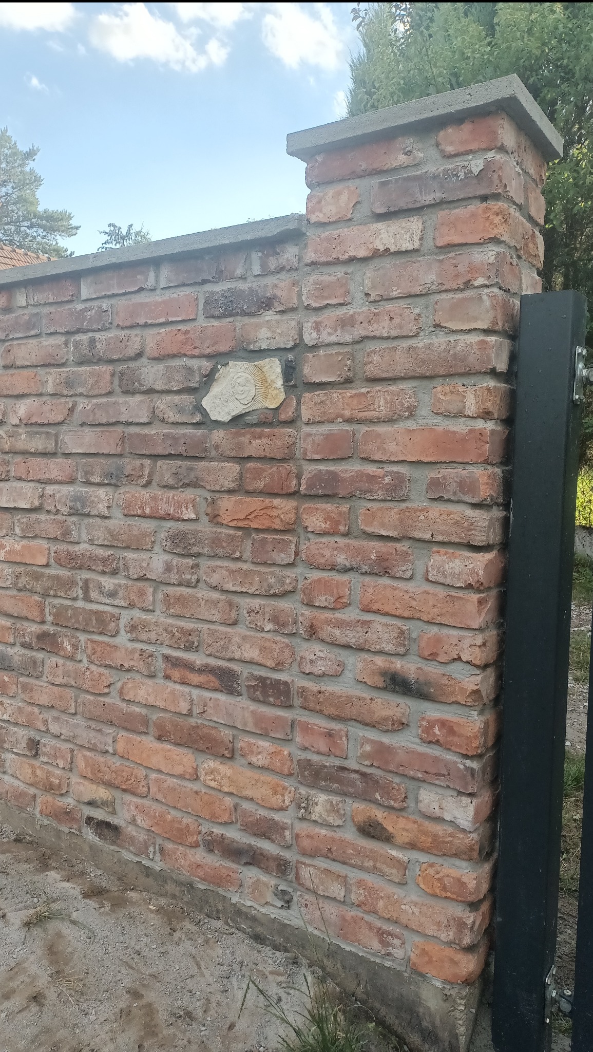 Murowany z cegły fragment ogrodzenia z betonowym wykończeniem i ozdobnym elementem. Widoczny fragment czarnej metalowej bramy. Ujęcie z dołu, w tle zieleń.