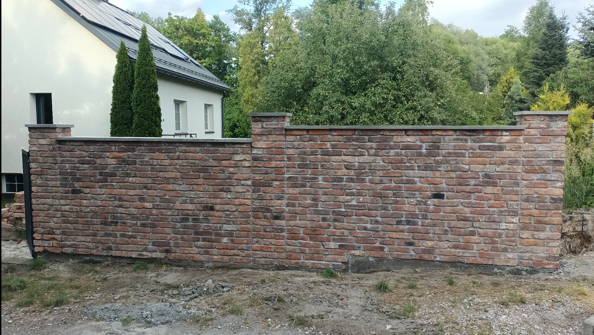 Mur z czerwonej cegły z szarymi elementami wykończeniowymi na tle domu z panelami słonecznymi i zieleni. Widoczne nierówności terenu i fragmenty gruzu.