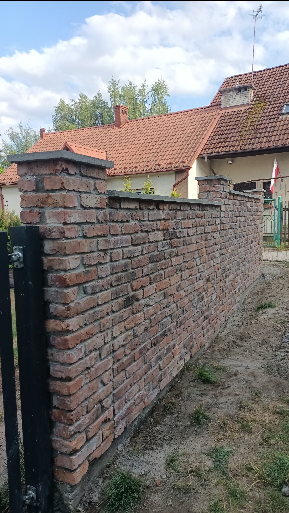 Murek z czerwonej cegły z szarymi elementami wykończeniowymi, oddzielający posesję. W tle dom z czerwoną dachówką i fragment metalowej furtki.