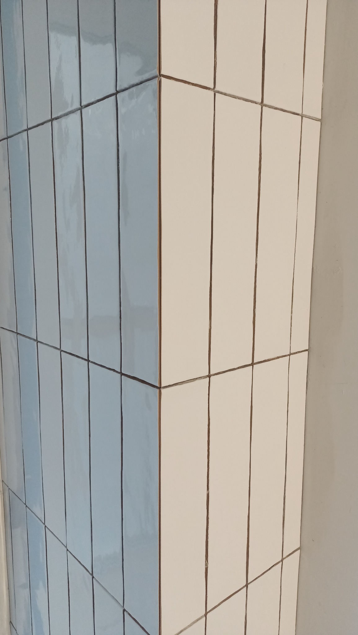 Ściana wyłożona wąskimi, pionowymi płytkami w dwóch kolorach: błękitnym i kremowym. Widoczny narożnik i fugi. Błyszcząca powierzchnia płytek odbija światło.