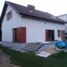 Dwukondygnacyjny dom z białą elewacją, brązowymi ramami okiennymi i drewnianymi okiennicami, widoczne schody wejściowe, niebieska betoniarka i stos piasku, sugerujące prace budowlane.