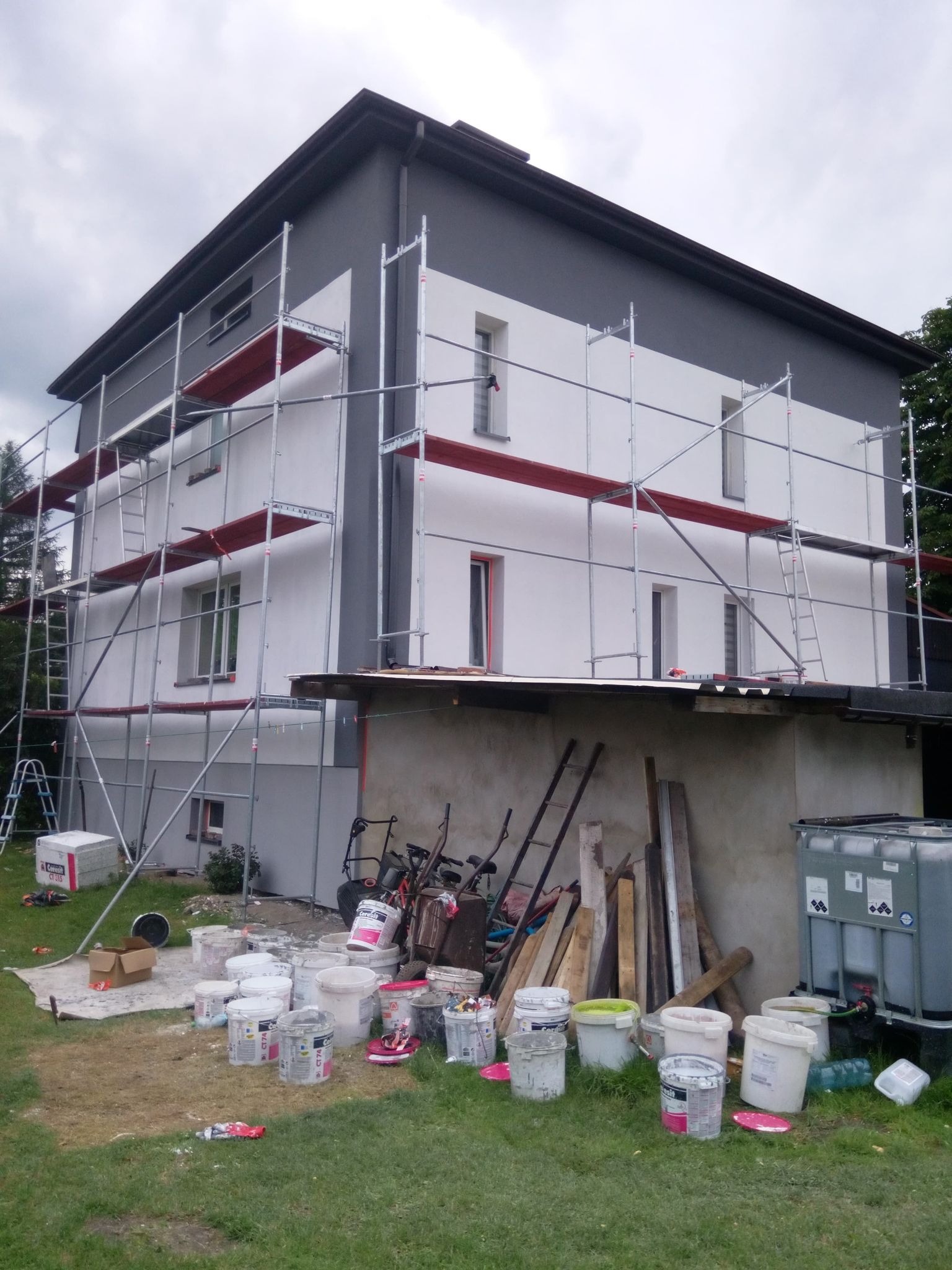 Dwukondygnacyjny dom w trakcie prac elewacyjnych: widoczne rusztowania, pomalowana część fasady w odcieniach szarości i bieli, liczne wiadra z farbą i narzędzia malarskie na trawniku.