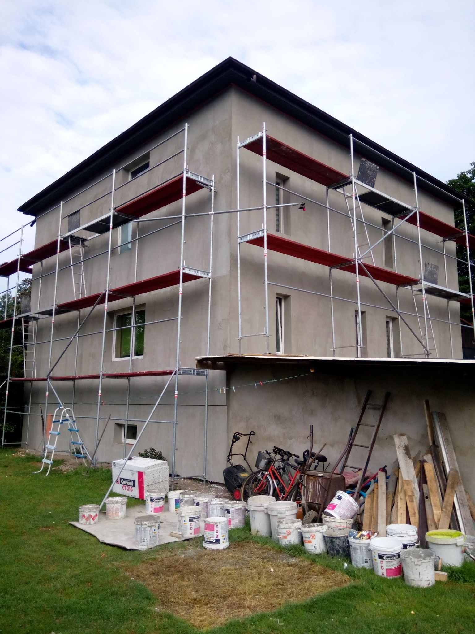 Dwupiętrowy dom w trakcie prac elewacyjnych, otoczony rusztowaniami, z widocznymi wiadrami farb i materiałów budowlanych na trawniku.