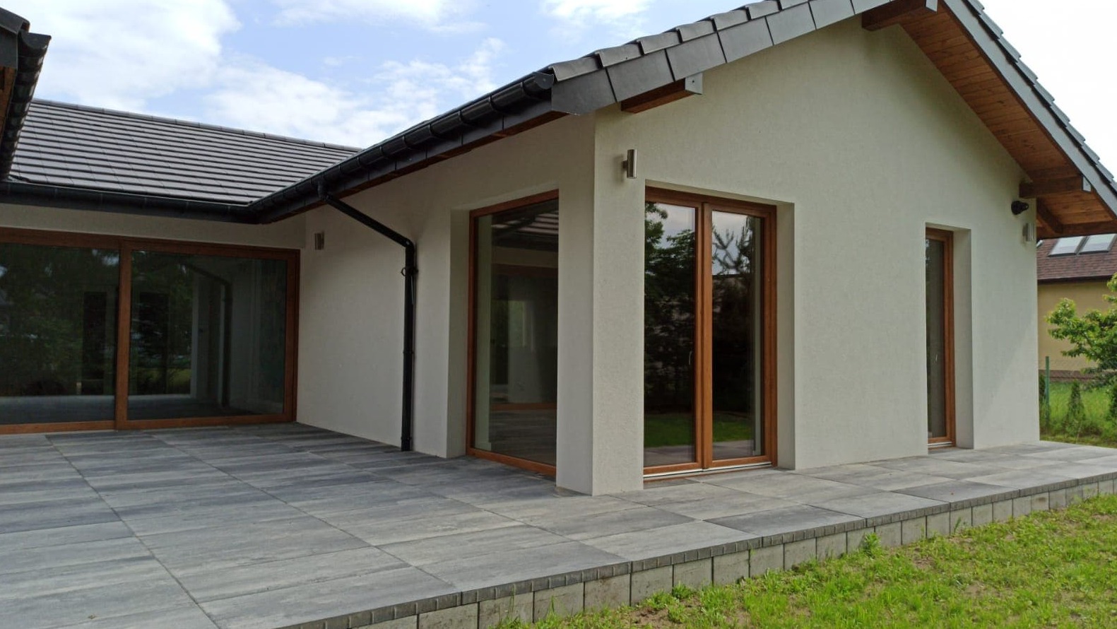 Dom jednorodzinny po wykonaniu elewacji w Lgota Górna, z widocznymi dużymi oknami z drewnianą ramą i szarą kostką brukową na tarasie.