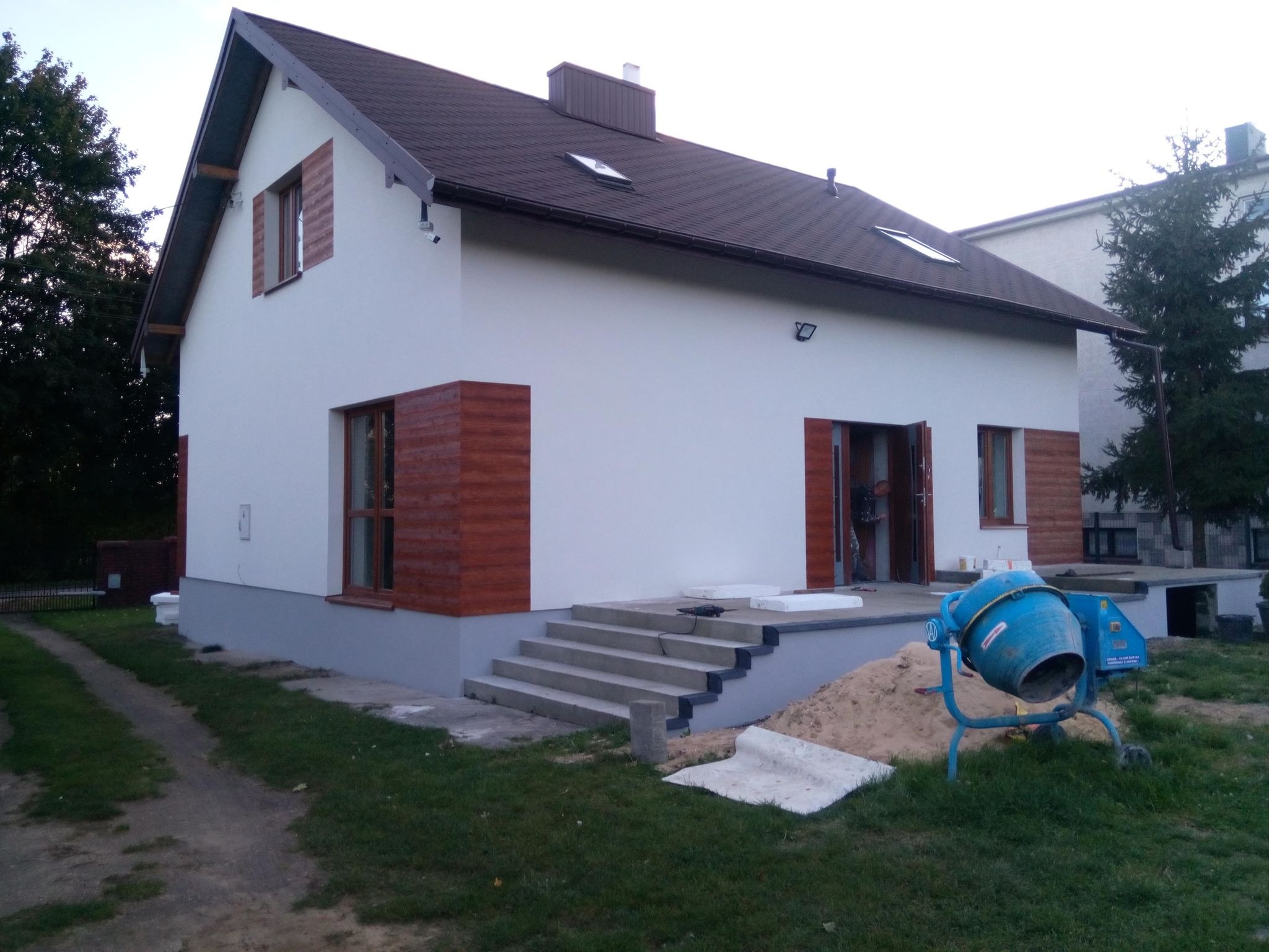 Dwukondygnacyjny dom z białą elewacją, brązowymi ramami okiennymi i drewnianymi okiennicami, widoczne schody wejściowe, niebieska betoniarka i stos piasku, sugerujące prace budowlane.