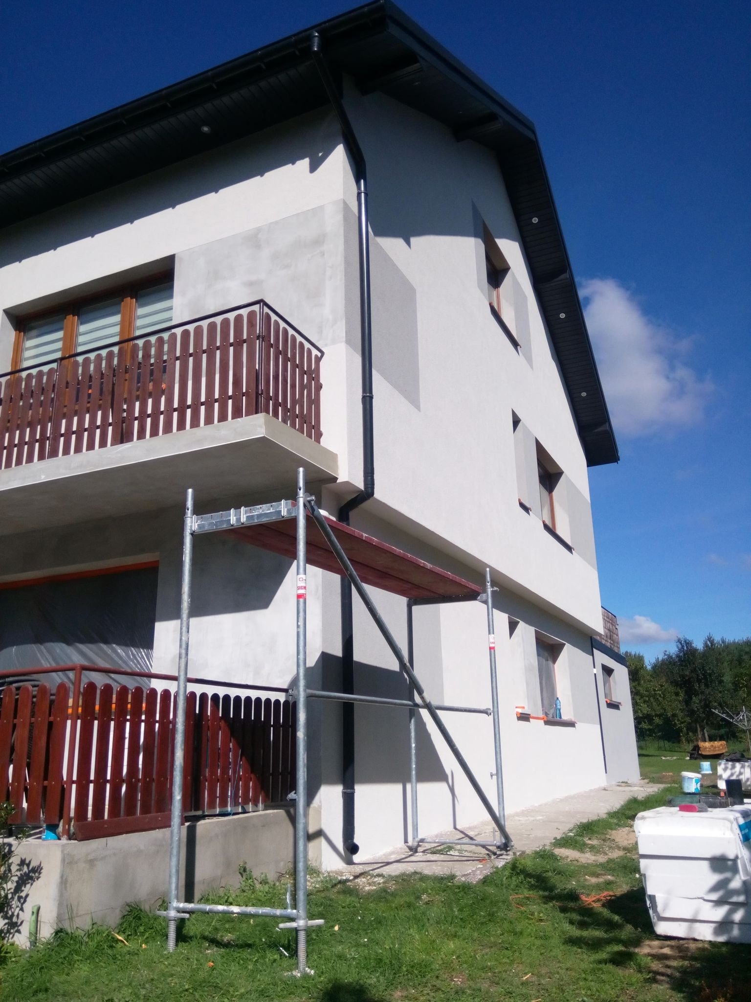 Dwupiętrowy dom z balkonem, na elewacji widoczne szare fragmenty, częściowo zakryty rusztowaniem, w tle błękitne niebo.