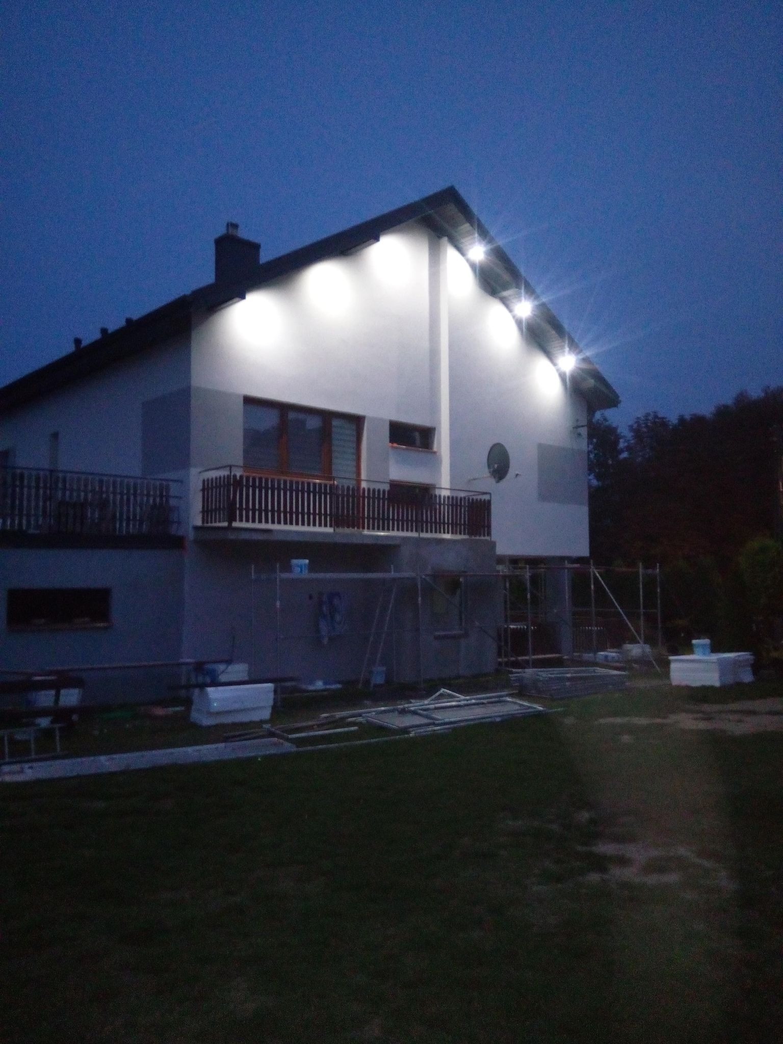 Dwupiętrowy dom jednorodzinny z balkonem oświetlony zewnętrznymi lampami halogenowymi w trakcie prac wykończeniowych, widoczne rusztowania i materiały budowlane na trawniku, zdjęcie zrobione...