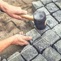 Ręczne układanie kostki granitowej z użyciem gumowego młotka i sznurka wyznaczającego linię, widok z góry na częściowo ułożoną nawierzchnię.