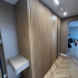 Usługi stolarskie - Nowoczesna szafa wnękowa z drewnianymi lamelami i wysuwaną półką, wkomponowana w aranżację przedpokoju z parkietem w jodełkę i oświetleniem sufitowym.