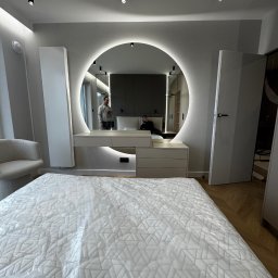Usługi stolarskie - Nowoczesna sypialnia z toaletką z okrągłym lustrem z podświetleniem LED. W lustrze odbija się stolarz podczas montażu. Minimalistyczny design, jasne kolory.