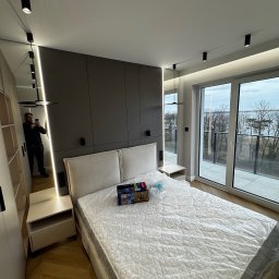 Usługi stolarskie - Nowoczesna sypialnia z łóżkiem, szafą wnękową z oświetleniem LED i dużym oknem balkonowym z widokiem na jezioro. W lustrze odbija się osoba robiąca zdjęcie.