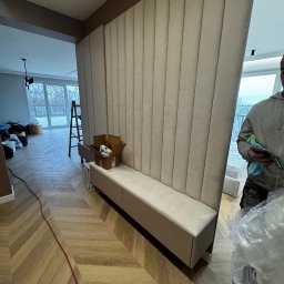 Usługi stolarskie - Tapicerowany panel ścienny z siedziskiem w przedpokoju, wzór jodełki na podłodze, karton z akcesoriami, drabina w tle, widok na okno. Montaż w trakcie.