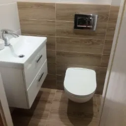 Nowoczesna, mała łazienka z wiszącą szafką umywalkową, toaletą podwieszaną z chromowanym przyciskiem spłukującym oraz płytkami imitującymi drewno na ścianie i podłodze.