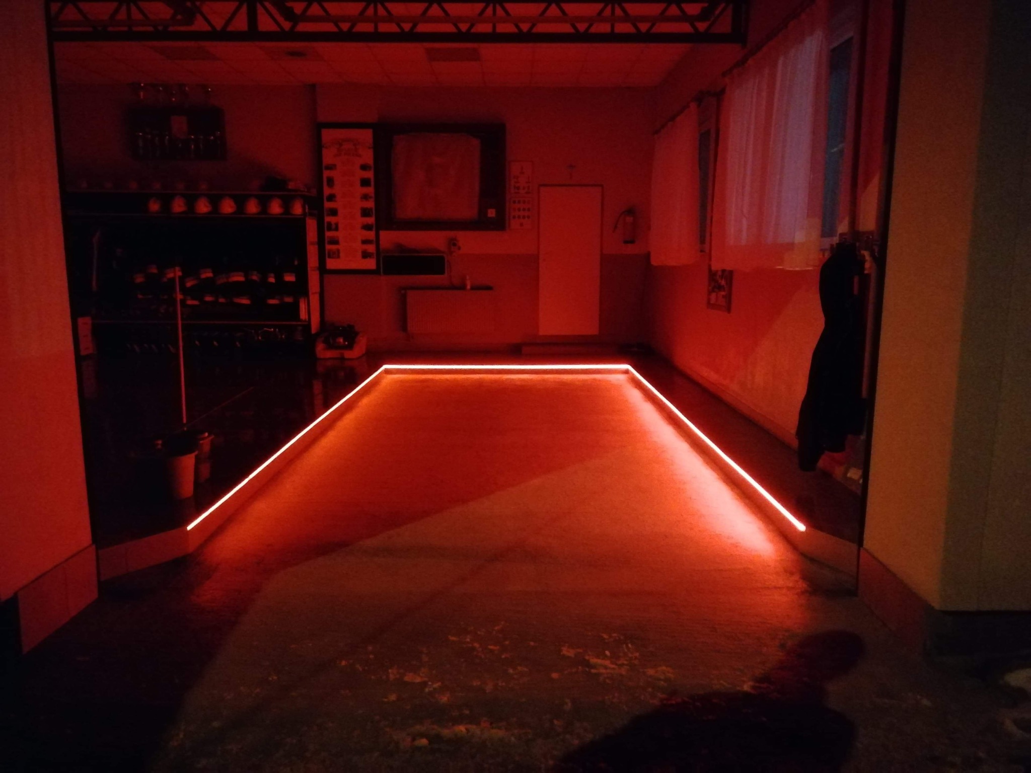 Garaż oświetlony czerwoną taśmą LED, tworzącą obrys podłogi, z widocznymi elementami wyposażenia i drzwiami w tle.