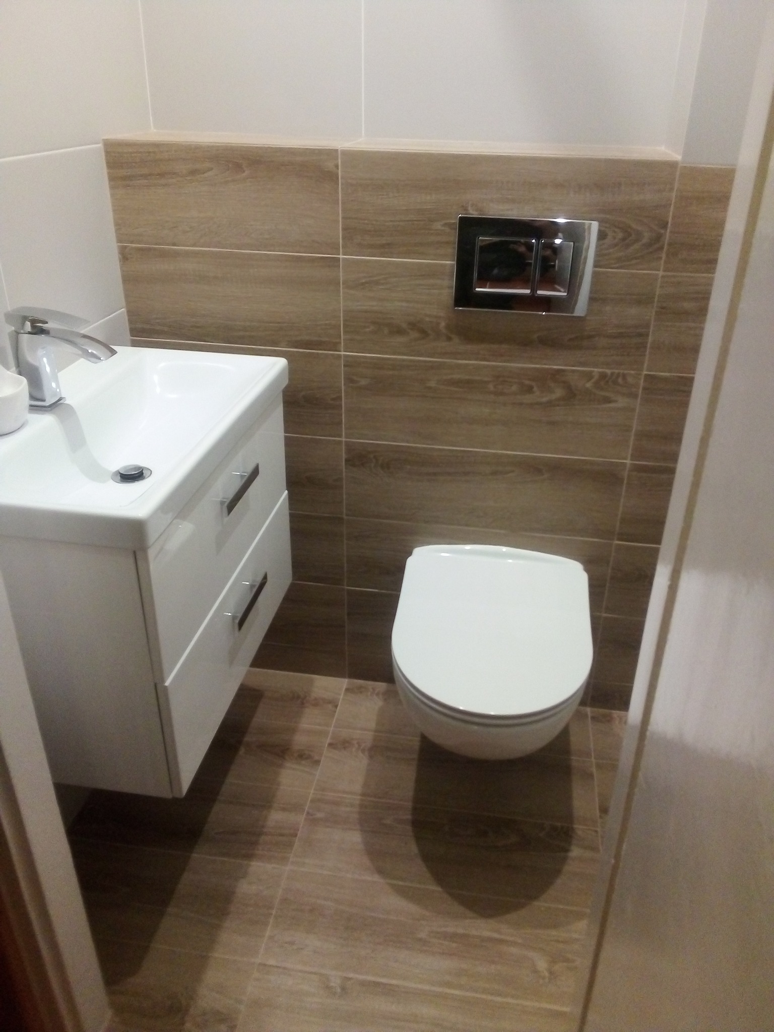 Nowoczesna, mała łazienka z wiszącą szafką umywalkową, toaletą podwieszaną z chromowanym przyciskiem spłukującym oraz płytkami imitującymi drewno na ścianie i podłodze.