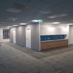 Tomasz Stenzel - Wnętrze biurowe po remoncie: szare ściany, białe drzwi, zabudowa kuchenna z niebieskim panelem, drabina malarska i wzorzysta wykładzina dywanowa.