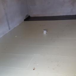 Tomasz Stenzel - Przygotowanie podłoża pod posadzkę: ułożone płyty izolacyjne na podłodze w surowym pomieszczeniu, widoczny odpływ podłogowy i fragment folii izolacyjnej przy ścianie.