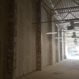 Tomasz Stenzel - Wnętrze hali w trakcie prac wykończeniowych, ściany po szpachlowaniu, widoczne elementy konstrukcji stalowej dachu, drabina i materiały budowlane.