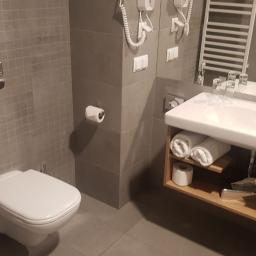 Tomasz Stenzel - Nowoczesna łazienka z szarymi płytkami, podwieszaną toaletą, umywalką z drewnianą szafką i zwiniętymi ręcznikami.