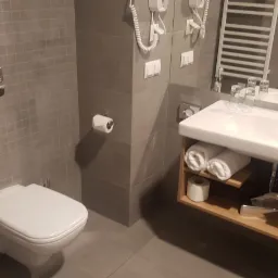 Nowoczesna łazienka z szarymi płytkami, podwieszaną toaletą, umywalką z drewnianą szafką i zwiniętymi ręcznikami.