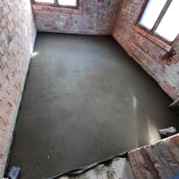 Tomasz Stenzel - Świeżo wylana, gładka wylewka samopoziomująca w pomieszczeniu z surowymi, ceglanymi ścianami i oknami w Tczewie. Widoczny fragment listwy dylatacyjnej przy ścianie.