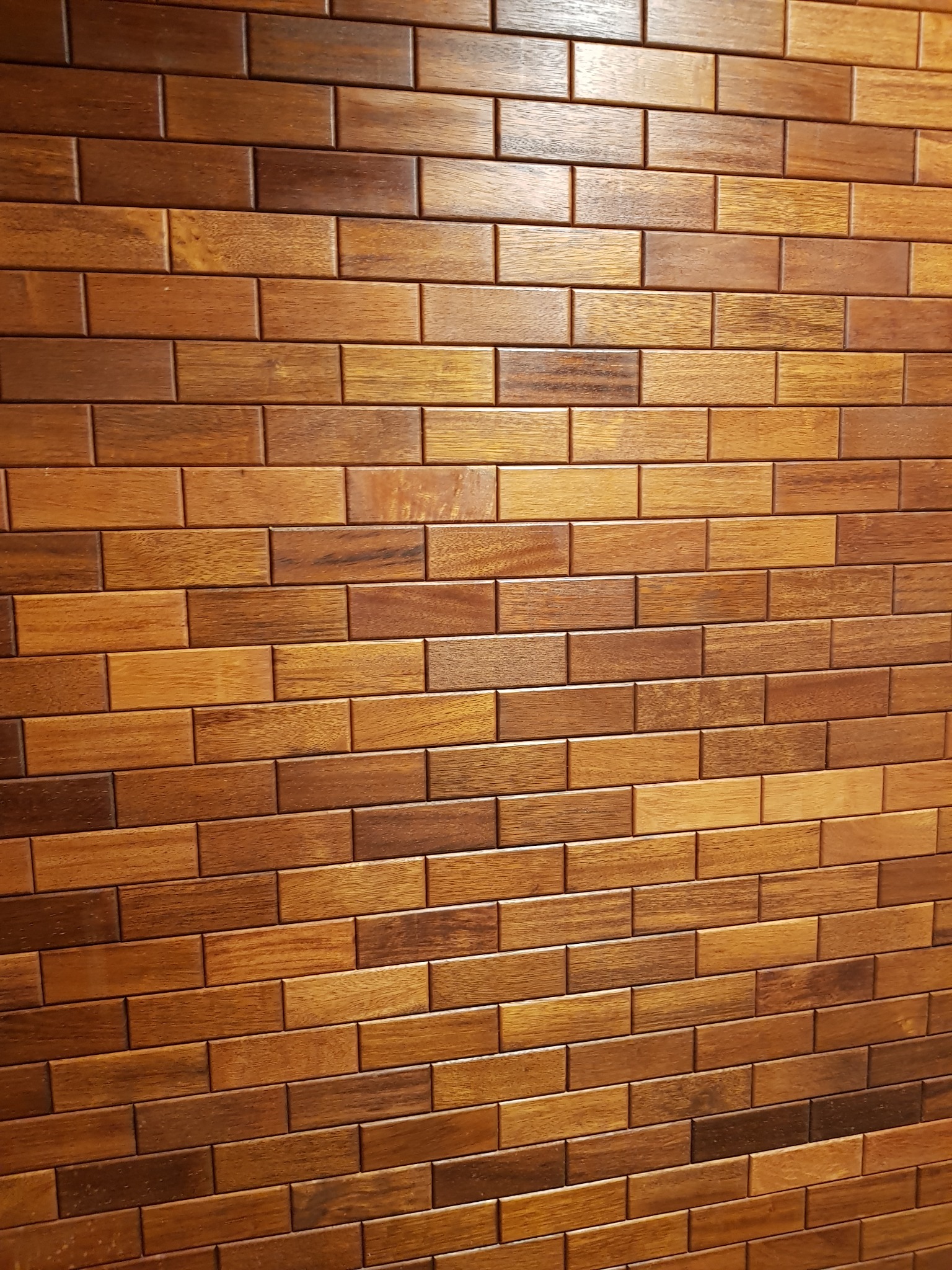 Ściana wyłożona błyszczącymi, drewnianymi płytkami w odcieniach brązu i beżu, ułożonymi w regularny wzór cegiełek.