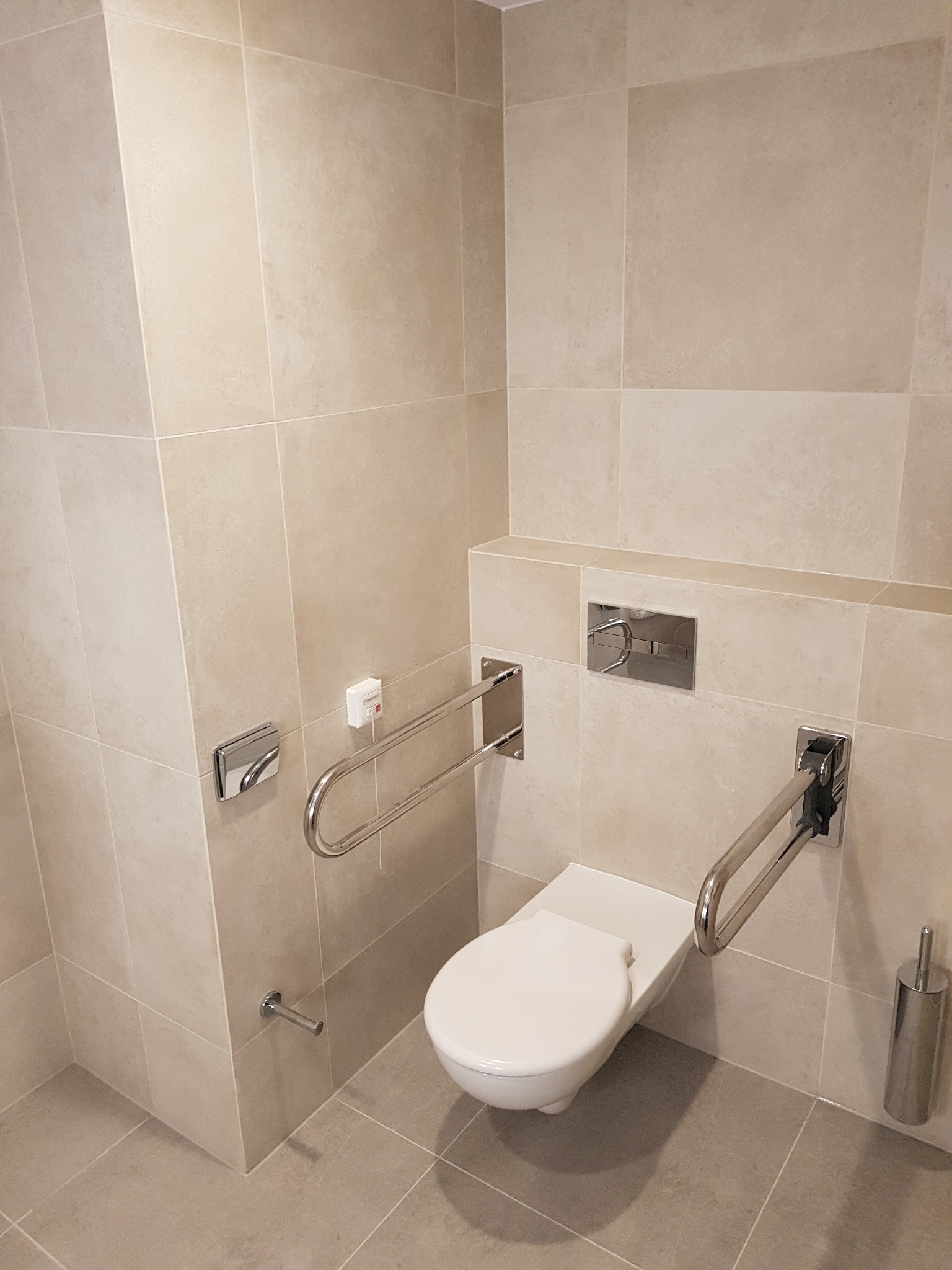 Beżowa łazienka z toaletą dla niepełnosprawnych, wyposażona w chromowane uchwyty, dozownik mydła i szczotkę do WC, wykończona płytkami w odcieniach beżu.