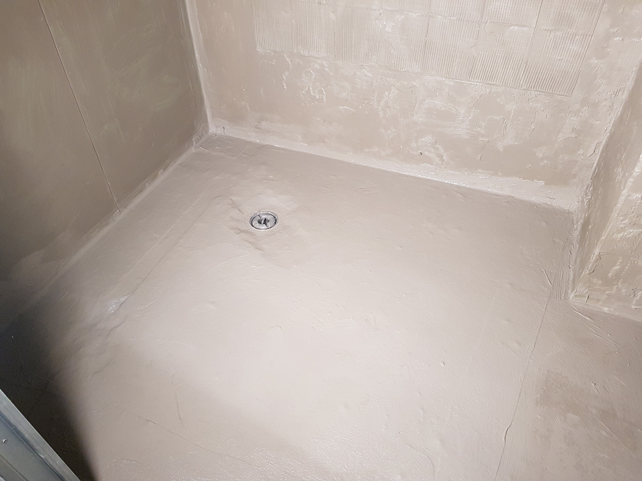 Przygotowana do położenia płytek podłoga i ściany w kabinie prysznicowej z odpływem liniowym, pokryte warstwą hydroizolacji w kolorze beżowym.