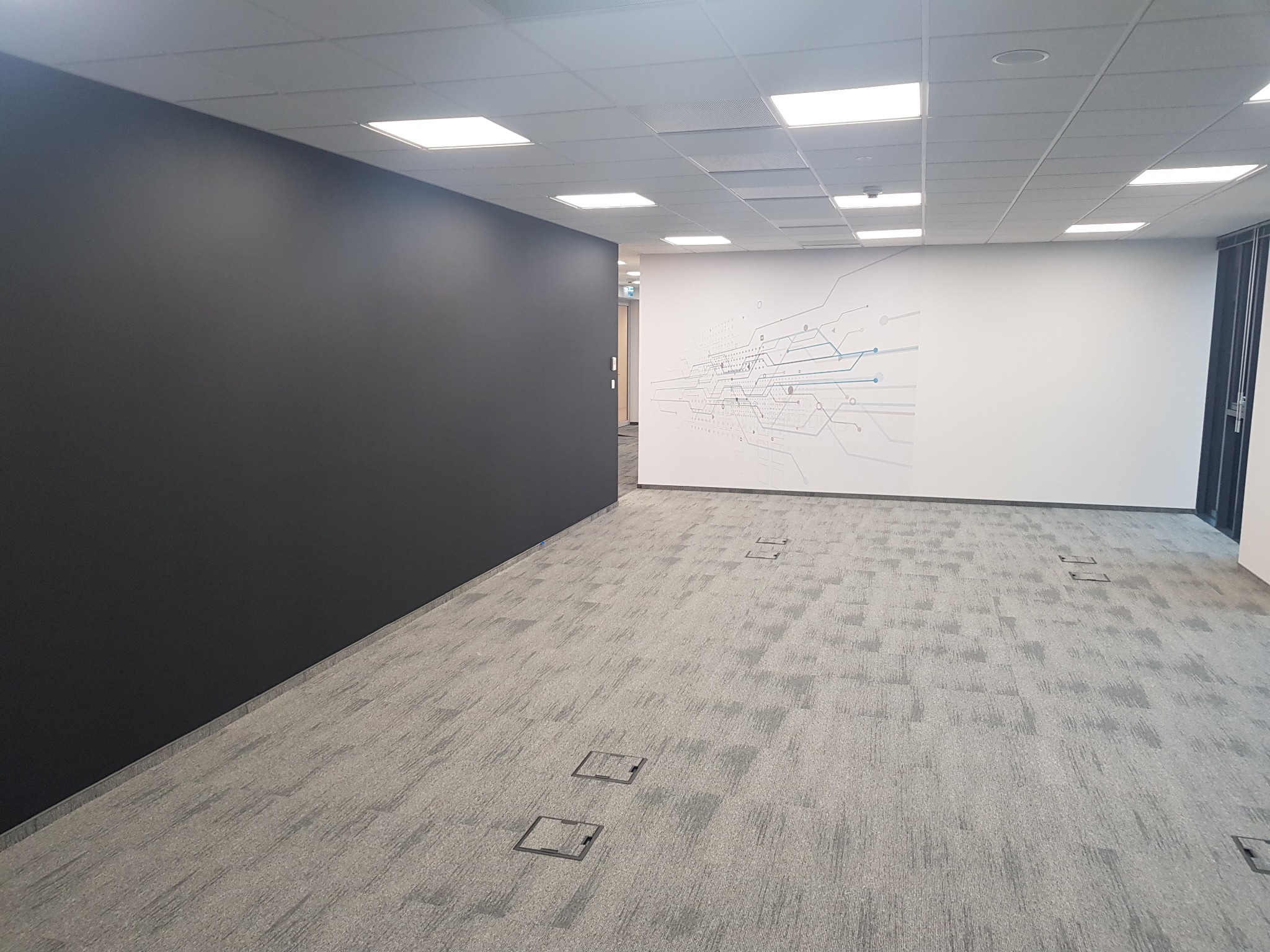 Wnętrze biurowe po remoncie: szara wykładzina dywanowa w kwadraty, czarna ściana po lewej, biała ściana z dekoracyjnym rysunkiem obwodu elektronicznego po prawej.