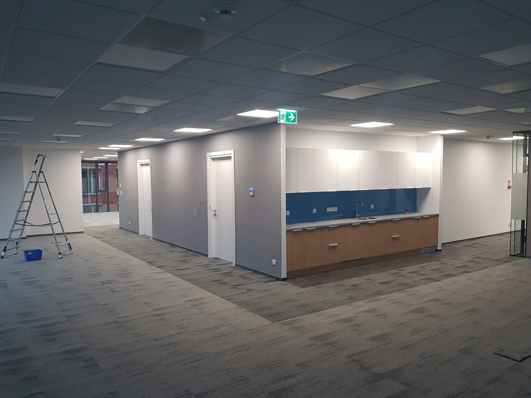 Wnętrze biurowe po remoncie: szare ściany, białe drzwi, zabudowa kuchenna z niebieskim panelem, drabina malarska i wzorzysta wykładzina dywanowa.