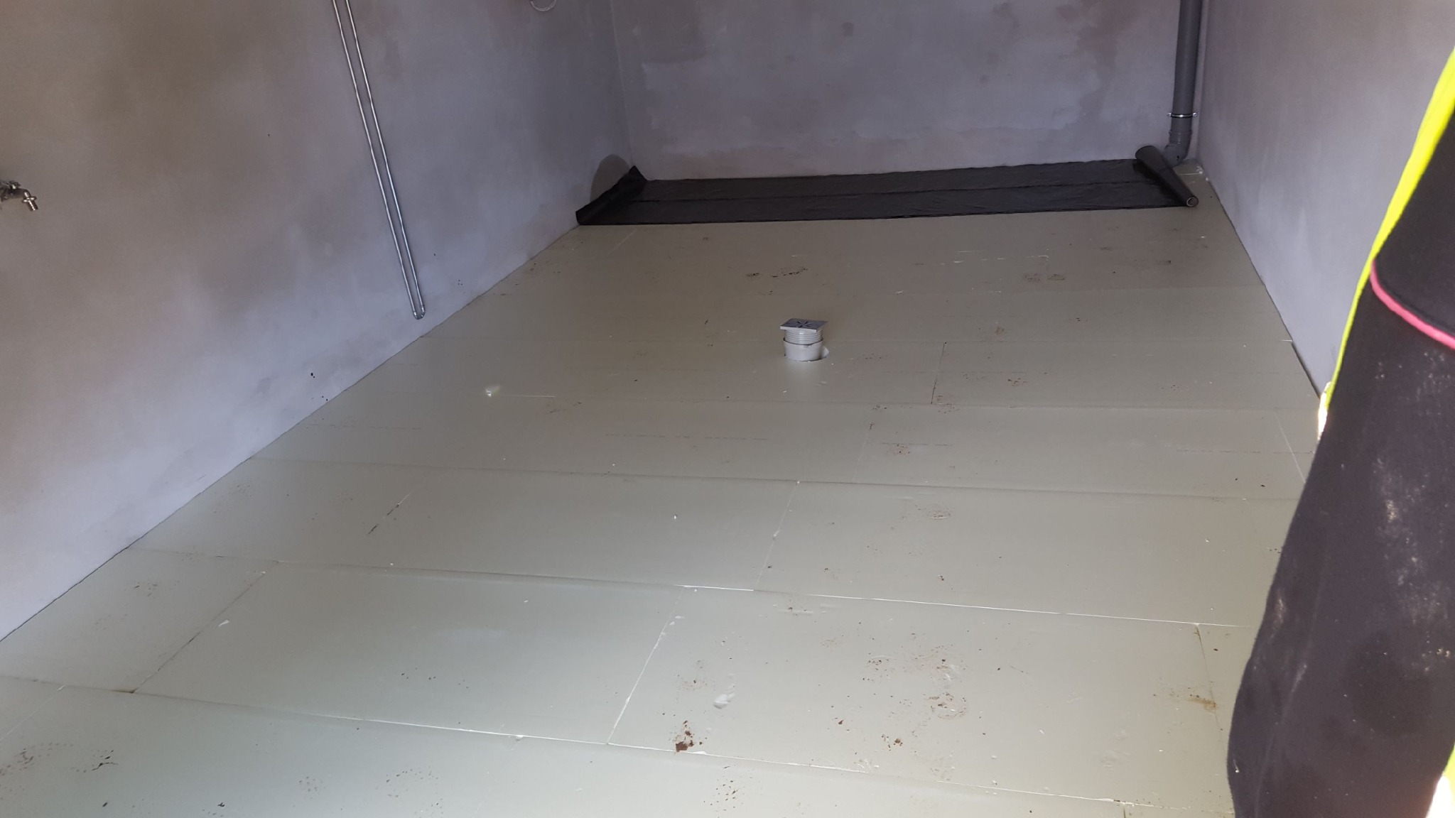 Przygotowanie podłoża pod posadzkę: ułożone płyty izolacyjne na podłodze w surowym pomieszczeniu, widoczny odpływ podłogowy i fragment folii izolacyjnej przy ścianie.