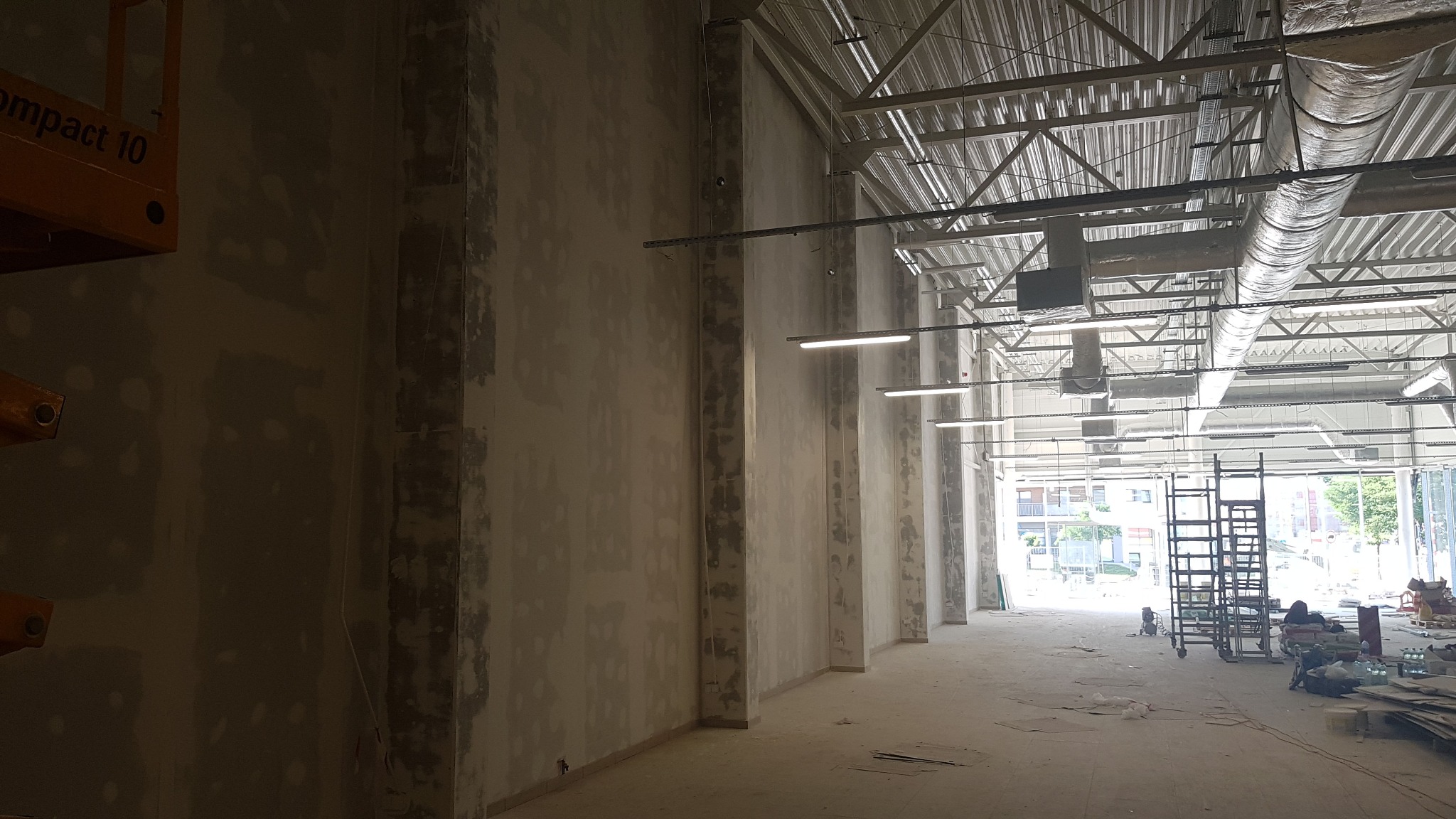 Wnętrze hali w trakcie prac wykończeniowych, ściany po szpachlowaniu, widoczne elementy konstrukcji stalowej dachu, drabina i materiały budowlane.