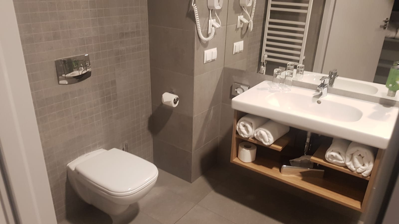 Nowoczesna łazienka z szarymi płytkami, podwieszaną toaletą, umywalką z drewnianą szafką i zwiniętymi ręcznikami.