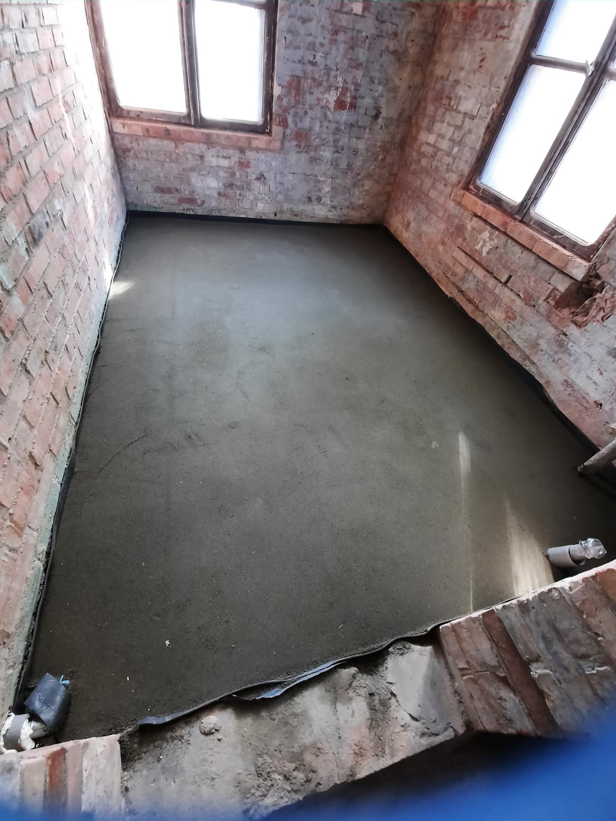 Świeżo wylana, gładka wylewka samopoziomująca w pomieszczeniu z surowymi, ceglanymi ścianami i oknami w Tczewie. Widoczny fragment listwy dylatacyjnej przy ścianie.