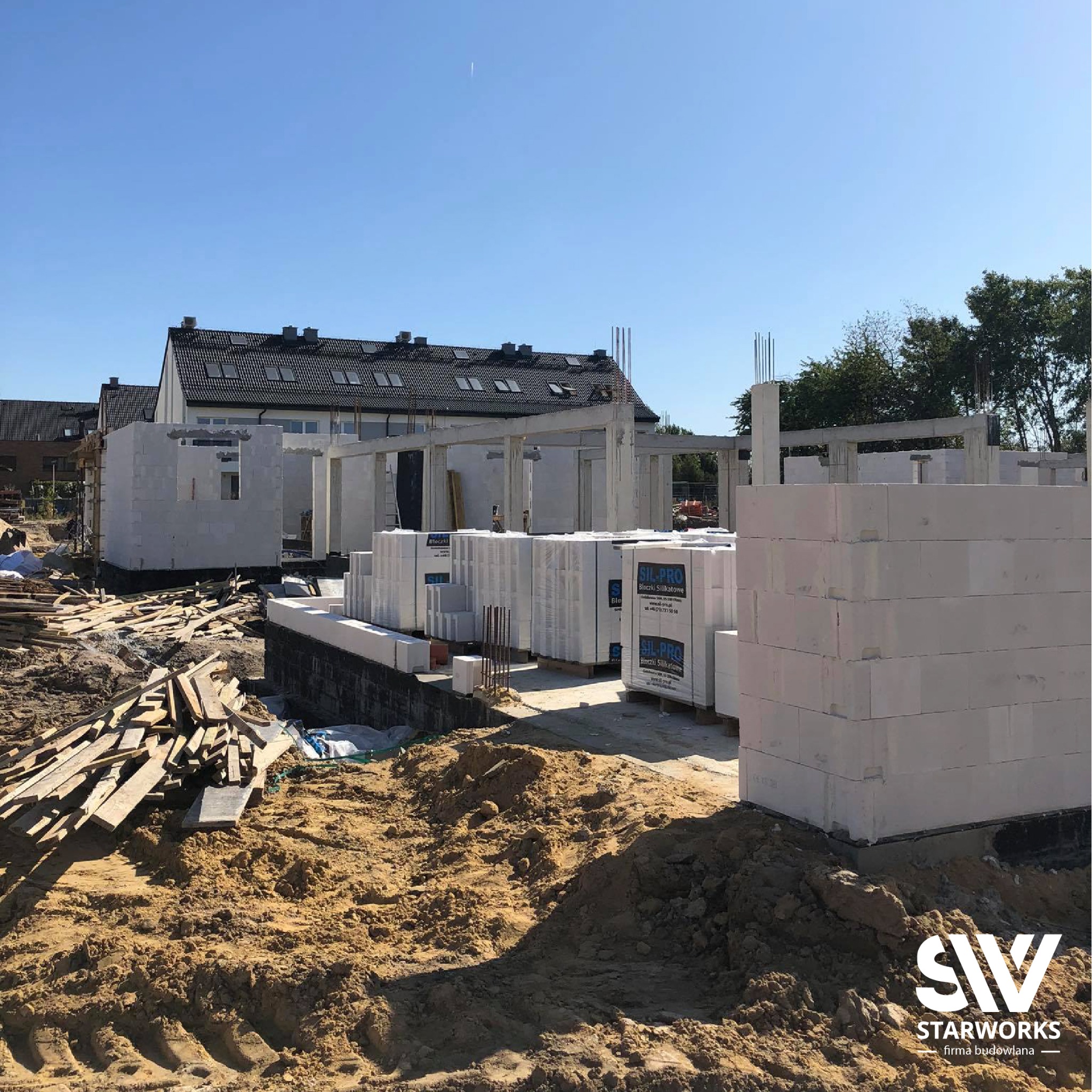 Budowa domu murowanego w trakcie realizacji: widoczne ściany z bloczków silikatowych, fundamenty, stosy materiałów budowlanych i porozrzucane deski. W tle budynki mieszkalne pod błękitnym niebem.