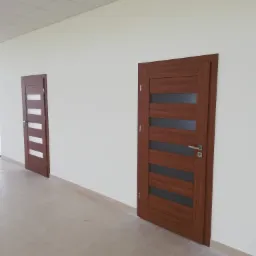 Dwie wewnętrzne, drewniane drzwi z przeszkleniami, w korytarzu z płytkami podłogowymi i oświetleniem sufitowym.